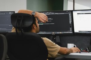 Un homme assis à un bureau, devant trois écrans d'ordinateur, concentré sur son travail.